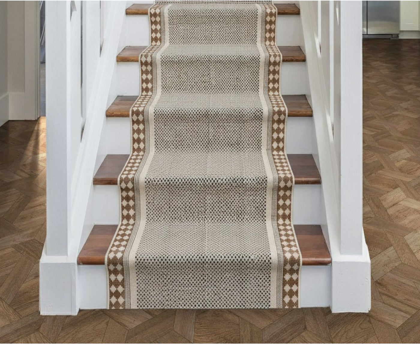 A woven stair runner with a textured beige and brown pattern and diamond borders, displayed on a staircase with wooden treads and white balusters.