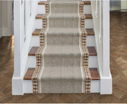 A woven stair runner with a textured beige and brown pattern and diamond borders, displayed on a staircase with wooden treads and white balusters.