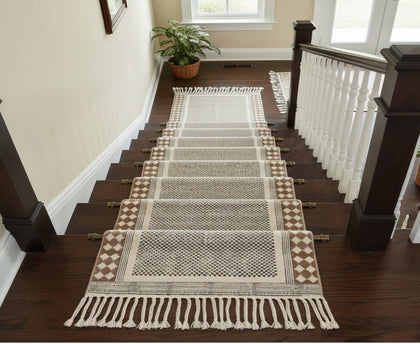 A long woven stair runner with fringe and geometric patterns in beige, brown, and gray, displayed on a dark wood staircase with white railings.
