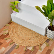 A semi-circular woven jute entry rug with tassels, placed on a wooden floor in front of white stairs, with potted plants nearby.