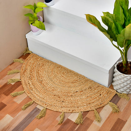 A semi-circular woven jute entry rug with tassels, placed on a wooden floor in front of white stairs, with potted plants nearby.