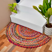 A colorful braided half-round entryway rug placed on a wood floor in front of white stairs, with potted plants nearby.