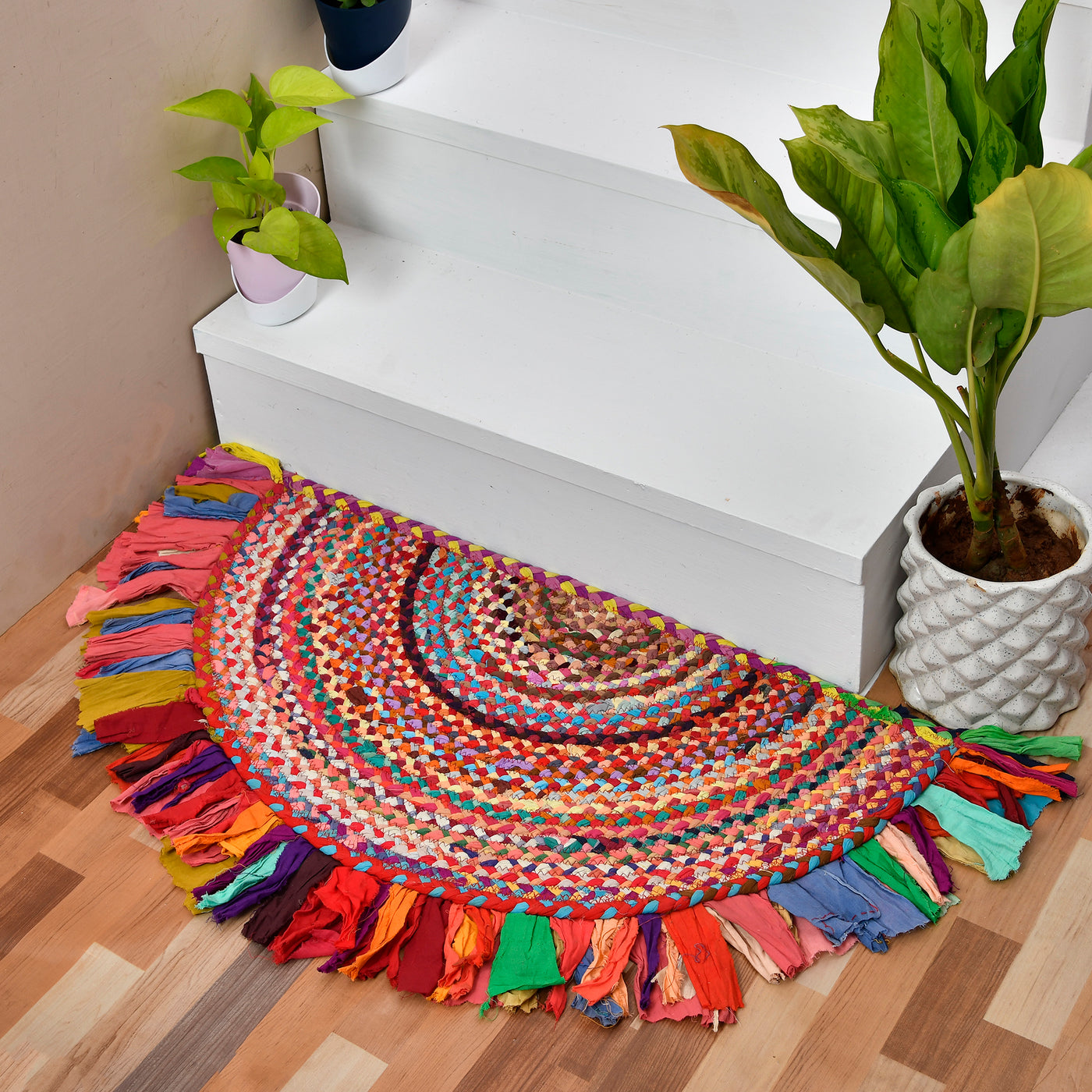 A colorful, semi-circular braided rug with fringe tassels, styled on a wooden floor next to white stairs and potted green plants.