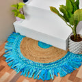 A round entryway rug with fringe, featuring a natural jute center and concentric blue woven bands, styled on a wooden floor next to white stairs and potted plants.