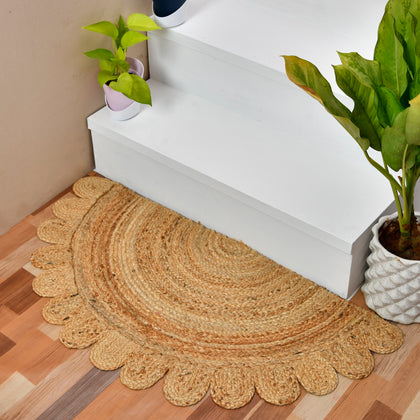 A round natural jute entry rug with a scalloped edge, placed on a wooden floor in front of white stairs, flanked by potted green plants.