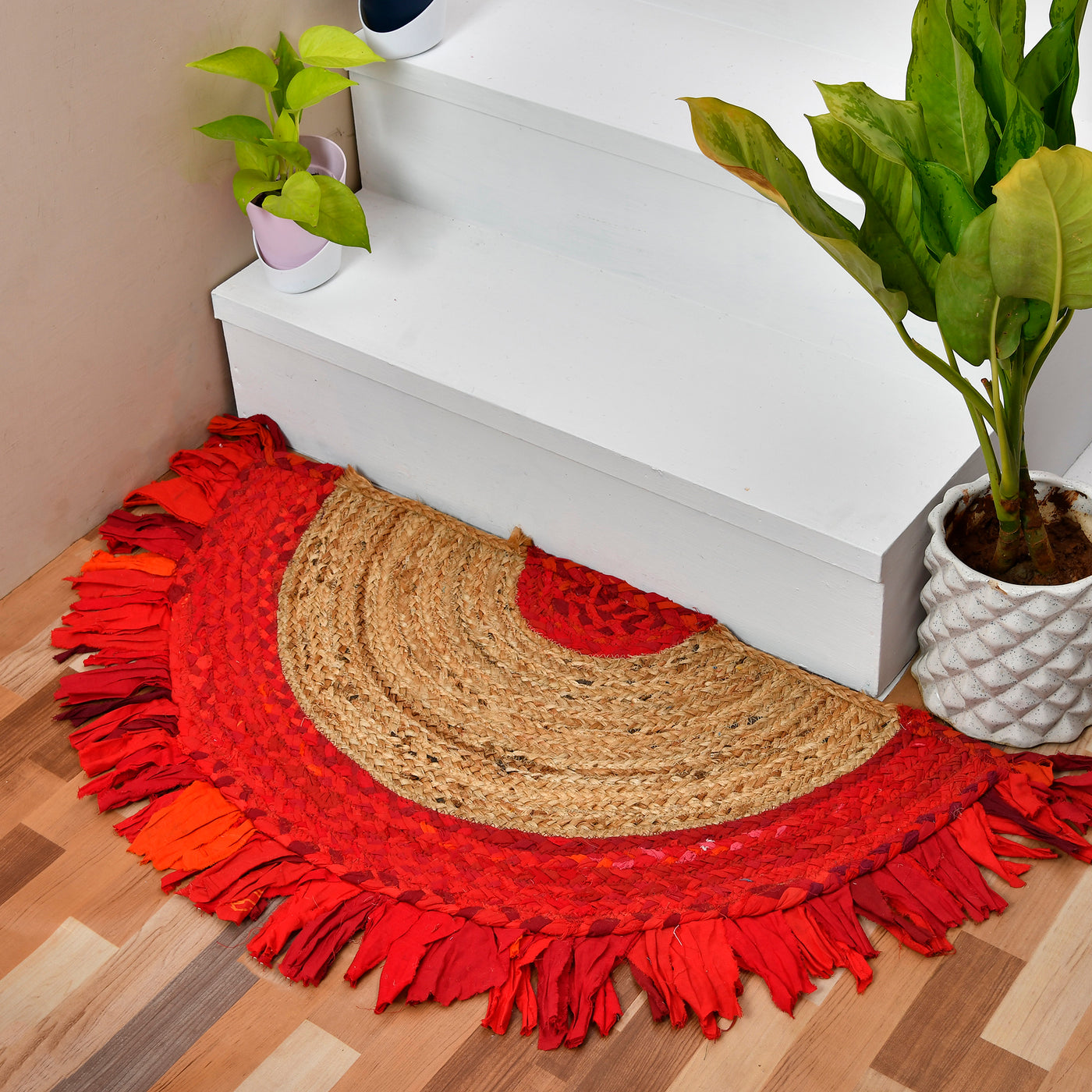 A round, semi-circular woven entryway rug with fringe, styled on a wooden floor next to white stairs and potted plants.