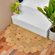 A round natural jute entryway rug with a scalloped edge, placed on a wooden floor in front of white stairs, styled with potted plants.